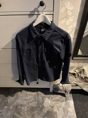 Mörkblå overshirt från Jack & Jones - Snygg mörkblå overshirt från Jack & Jones Core med klassisk krage och två bröstfickor. Overshirten har knappar framtill och är tillverkad i ett mjukt material som ger en avslappnad look. Perfekt att bära över en t-shirt för en casual stil.