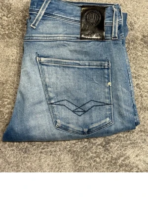 Replay ljusblå jeans med raka ben - Säljer ett par ljusblå jeans från Replay med raka ben och klassisk femficksmodell. Jeansen har snygga slitningar och Replay-logga på bakfickan samt svart patch bak i midjan. Perfekta till en avslappnad stil. Storlek 30/32
