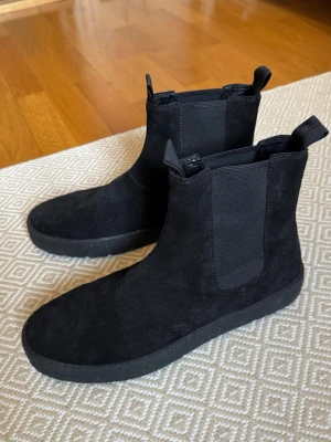 Svarta chelsea boots i mocka - Ett par nygga svarta chelsea boots i mocka från Vagabond. Bootsens design är stilren med rund tå och platt sula. Dom är i mycket bra skick, bara använda ett par gånger. Tveka inte att skriva om du ha några funderingar!