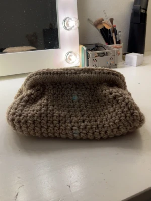 Beige stickad kuvertväska - Säljer en handgjord beige/brun kuvertväska i grovt stickat material. Väskan har en mjuk och chunky känsla med enkel design, perfekt för dig som gillar unika accessoarer. Ingen synlig logga eller märke. Skulle säga att väskan är 9 gånger 17 I metallställningen 💗