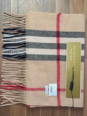 Burberry Halsduk - Klassisk beige halsduk från Burberry i 100% kashmir med det ikoniska rutmönstret i svart, vitt och rött. Halsduken har fransar i ändarna och känns både lyxig och tidlös. Perfekt accessoar för att lyfta vilken outfit som helst. Halsduken är oanvänd (ny). Skriv vid minsta lilla fundering!
