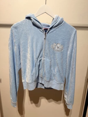 Ljusblå velour hoodie Juicy Couture - Säljer en ljusblå zip-up hoodie från Juicy Couture i mjuk velour. Hoodien har huva, fickor fram och ikonisk Juicy Couture-logga med glitterdetaljer både fram och bak. Perfekt för dig som gillar en avslappnad men ändå trendig stil.
