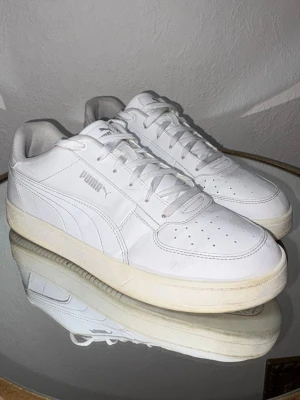 Vita sneakers från Puma - Stilrena vita sneakers från Puma med klassisk låg profil och rund tå. Skorna har snörning, perforerade detaljer på tån och är tillverkade i syntetmaterial med en vit sula. Perfekta för en clean och sportig look.