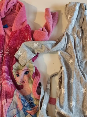 Frost morgonrock & grå stjärnrock stl 110/116 - Två mysiga morgonrockar för tonåringar: en rosa med Frost-motiv och texten 'Winter Magic', samt en grå med vita stjärnor. Båda har huva och knytskärp i midjan. Perfekta för lata morgnar eller kvällar hemma.