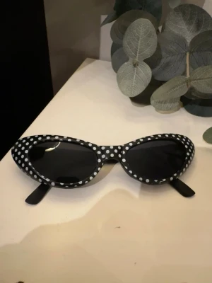 Svarta solglasögon med vita prickar - Coola svarta solglasögon med vita prickar och cat eye-form. Bågarna är i plast och har en retroinspirerad look som sticker ut. Perfekta för dig som vill ha en lekfull och trendig accessoar till din outfit.