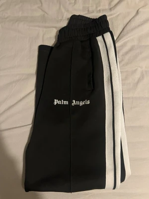Palm Angels byxor - Svarta mjukisbyxor från Palm Angels med vita sidoränder och logga på vänster ben. Byxorna har elastisk midja och raka ben, perfekta för en chill och sportig look. Storlek M, priset är inte hugget i sten!!! 