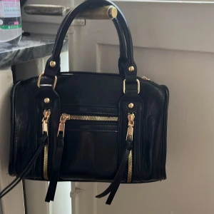 Svart handväska med gulddetaljer - Snygg svart handväska i skinnimitation med guldfärgade dragkedjor och detaljer. Väskan har två rejäla handtag, flera fack med dragkedja framtill och en stilren, boxig form. Perfekt storlek för att få plats med allt du behöver. Enkel och klassisk design som passar till många outfits.Köpte väskan på plick så vet inte märket. 