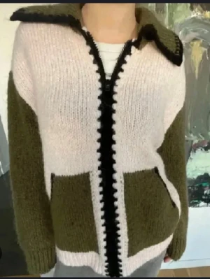 Stickad kofta i olivgrönt och vitt - Mysig stickad kofta med olivgröna ärmar och fickor, vit kropp och svarta detaljer längs dragkedjan och kragen. Koftan har en avslappnad passform, stora fickor framtill och ribbstickade muddar. Perfekt för lager på lager och chill stil.