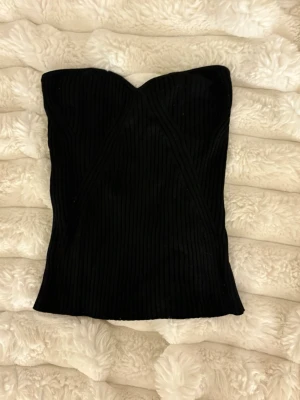 Svart ribbad tubtopp från Lindex - Snygg svart ribbad tubtopp från Lindex. Toppen är axelbandslös och har en tight passform med markerad byst. Perfekt att styla med jeans eller kjol för en trendig look.