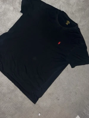 Svart t-shirt från Ralph Lauren - Klassisk svart t-shirt från Ralph Lauren med rund halsringning och röd broderad logga på bröstet. Enkel och stilren design som passar till allt. Tillverkad i mjuk bomull för skön känsla hela dagen. Kommer tvättas innan skickning