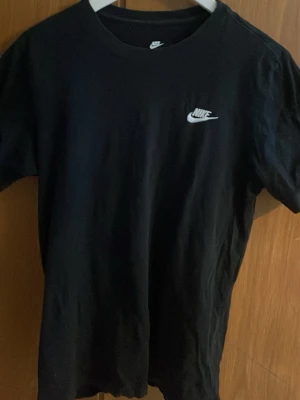 Svart Nike T-shirt med logga - Klassisk svart T-shirt från Nike med vit logga på bröstet. T-shirten har rund halsringning och korta ärmar. Tillverkad i mjuk bomull som känns skön mot huden. Perfekt för en clean och sportig look.