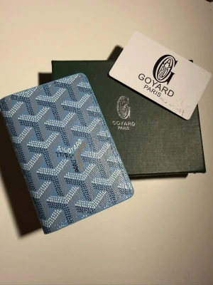 Blå Goyard plånbok med mönster - Säljer en snygg blå plånbok från Goyard med det klassiska grafiska mönstret i vitt och ljusblått. Plånboken är kompakt och har en lyxig känsla, perfekt för kort och sedlar. Kommer med originalask och autenticitetskort.box medföljs ej
