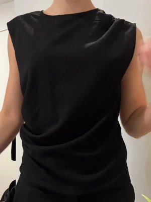 Svart ärmlös topp med drapering - Snygg svart ärmlös topp från HM med diskret drapering på ena sidan och på ena axeln. Använd ett fåtal gånger
