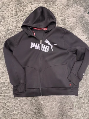 Svart hoodie med dragkedja från Puma - Snygg svart hoodie från Puma med dragkedja framtill och stor logga i vitt på bröstet. Klassisk huva och två fickor fram. Tillverkad i mjukt material som känns skönt mot huden. Perfekt för en sportig och avslappnad stil.