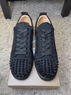 Svarta sneakers med nitar och röd sula - Säljer ett par svarta sneakers i mocka med coola svarta nitar på tån och klassisk röd sula. Skorna har snörning och en stilren, modern look som sticker ut. Perfekta för dig som vill ha något extra till outfiten.