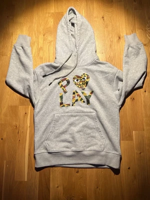 Comme des Garçons hoodie med camomönster - Snygg grå hoodie från Comme des Garçons Play med stor PLAY-logga och hjärta på bröstet i camomönster. Klassisk känguruficka framtill och justerbar huva med dragsko. Perfekt för dig som gillar streetwear och vill sticka ut.