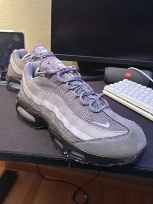 Nike Air Max 95 grå sneakers - Nike Air Max 95 sneakers i olika nyanser av grått med mesh och syntetmaterial. Klassisk vågig design, synlig Air-dämpning i sulan och snörning framtill. Perfekta för dig som gillar streetwear och sportig stil.