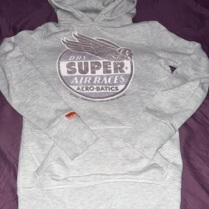 Grå Superdry hoodie med  tryck  - Snygg ljusgrå hoodie från Superdry i storlek XS. Tröjan har ett stort vintageinspirerat tryck på bröstet med texten 'Super Air Races Aero-Batics' och en cool vinge. Klassisk känguruficka och huva med dragsko. Perfekt för chill dagar. Köpt för 699 säljer för 499 men kan gå ner i pris förstås 