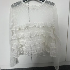 Vit prickig blus med volanger Gina Tricot - Supersöt vit blus från Gina Tricot i storlek XS. Blusen är transparent med prickigt mönster och har flera lager volanger framtill. Långärmad och tillverkad i ett lätt syntetmaterial som ger en luftig känsla. Perfekt för dig som gillar romantiska detaljer och unik stil.