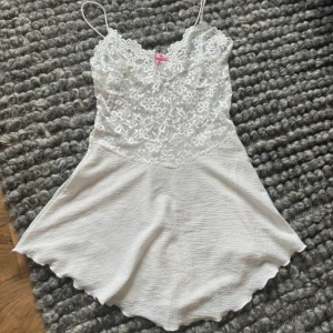 Vit spetsklänning med smala axelband - Supersöt vit kortklänning med smala axelband och blommig spets upptill. Kjolen är lätt och har vågig kant. Perfekt för sommarens varma dagar eller studentfirande. Spetsen ger en romantisk vibe och den lilla rosetten framtill är en gullig detalj.
