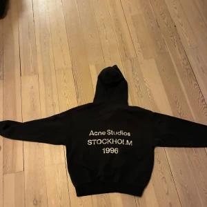 Svart hoodie Acne Studios Stockholm - Svart hoodie från Acne Studios med stor huva och tryck på ryggen: 'Acne Studios STOCKHOLM 1996'. Klassisk loose fit och ribbade muddar. Perfekt för dig som gillar stilrena streetwear-plagg med svensk design.