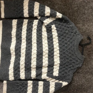Gråvit stickad randig tröja - Säljer en mysig stickad tröja med breda grå och vita ränder. Tröjan har ett klassiskt kabelstickat mönster och rund hals. Perfekt för dig som gillar en chill och stilren look under kyliga dagar.