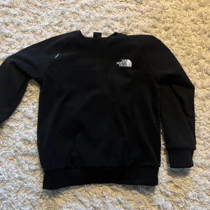 Svart tröja från The North Face - Säljer en stilren svart tröja från The North Face. Tröjan har en klassisk design med långärmade raglanärmar och en rund halsringning. Perfekt för en avslappnad look. Logotypen är tryckt på bröstet i vitt. 