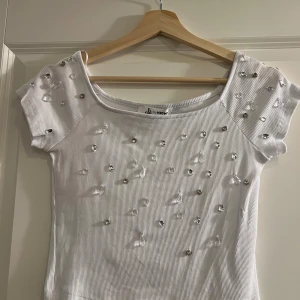 Off-shoulder topp med pärlor  - Säljer en vit ribbad topp från Olé by Koton i storlek M. Toppen är off-shoulder och är dekorerad med glittrande pärlor och stenar på framsidan och axlarna. Materialet är mjukt och stretchigt, perfekt för dig som gillar detaljer och vill sticka ut. Helt oanvänd!