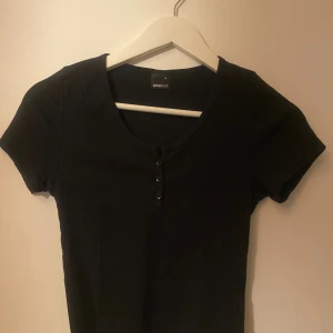 Svart ribbad topp från Gina Tricot - Svart ribbad topp från Gina Tricot med korta ärmar och rund halsringning. Toppen har tre knappar framtill som detalj och är gjord i mjuk bomull. Perfekt att styla med jeans eller kjol för en chill look.