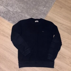 Svart sweatshirt från Lacoste - En stilren svart sweatshirt från Lacoste med svart logo på bröstet. Tröjan har rund hals, långa ärmar och ribbade muddar vid ärmslut och nederkant. Perfekt för dig som gillar klassisk och enkel stil.
