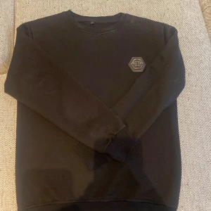 Svart sweatshirt från Philipp Plein - Svart sweatshirt från Philipp Plein med rund hals och ribbade muddar. På bröstet sitter en snygg hexagonformad logga i svart och silver. Tröjan har en clean look och är perfekt för dig som gillar stilrena plagg med exklusiv känsla.