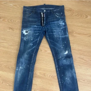 Dsquared2 Cool Guy jeans blå stl 46 - Snygga Dsquared2 Cool Guy jeans i mörkblå tvätt med slitningar och färgstänk för en edgy look. Modellen har smal passform, klassiska fem fickor och dragkedja. Tillverkade i bomull med en skön stretch. Perfekta för dig som gillar streetstyle.