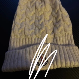 Vit stickad Moncler mössa med pälsboll - Snygg vit mössa från Moncler med grovstickat flätmönster och bred ribbad kant. På toppen sitter en fluffig vit pälsboll som ger extra stilpoäng. Märkeslogga framtill. Perfekt för dig som vill ha en varm och trendig mössa i vinter.