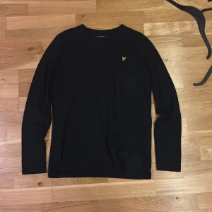 Svart långärmad tröja Lyle & Scott - Svart långärmad tröja från Lyle & Scott med klassisk rund hals och gul broderad logotyp på bröstet. Tröjan är tunn och gjord i mjuk bomull och har en stilren, enkel design som passar perfekt till jeans eller chinos. Snygg och tidlös för dig som gillar cleana plagg.