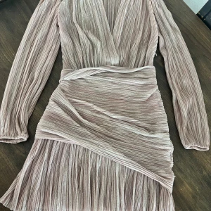 Glittrig kortklänning från Maje - Superfin glittrig kortklänning från Maje i ljus champagnefärg med plisserat tyg och omlottkänsla. Klänningen har långa ärmar, v-ringning och markerad midja. Perfekt för dig som vill sticka ut med en elegant och trendig look.