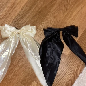 Stora satinrosetter i svart och vitt - Två stora rosetter i glansigt satinmaterial, en i svart och en i vit. Båda har långa band och fästs enkelt med metallspänne baktill. Perfekt för att ge en extra touch till din frisyr och funkar lika bra till utsläppt hår som till en hästsvans. Knappt använda. Skriv till mig innan du köper!