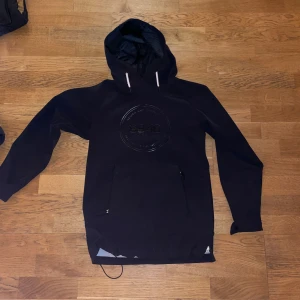 Svart skidjacka från 8848 Altitude - Svart skidjacka från 8848 Altitude med stor logga på bröstet, huva med vita snören och praktisk magficka med dragkedja. Jackan har lång ärm och är tillverkad i slitstark syntet med mjuk fleece på insidan. Perfekt för dig som gillar att vara ute när det är kallt och snöigt. Storlek 160. Skick 9/10