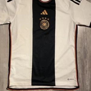 Tysklands fotbollströja Adidas M - Säljer en officiell Tysklands fotbollströja från Adidas, modell 2022/2023. Tröjan är vit med ett bred svart vertikalt fält i mitten, guldiga detaljer och Tysklands landslagsemblem med fyra stjärnor ovanför. Klassisk Adidas-design med tre svarta ränder på axlarna.  Storlek: M (Medium) Modell: Adidas Aeroready – andas och transporterar bort fukt Skick: Mycket bra / knappt använd Färger: Vit, svart, guld, röd och mörkbrun kantlinje