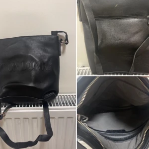 Svart axelväska från Don Donna - Snygg svart axelväska från Don Donna i skinn med präglad logga på framsidan. Väskan har justerbar axelrem, dragkedja upptill och en praktisk ytterficka med dragkedja. Perfekt storlek för vardagsgrejer och har ett stilrent, klassiskt utseende.