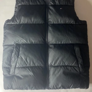 Svart dunväst från Tommy Hilfiger - Svart, quiltad dunväst från Tommy Hilfiger med diskret logga på bröstet och stor logotyp på insidan. Västen har dragkedja framtill och två sidofickor. Materialet är polyester och designen är stilren med breda, vadderade paneler.