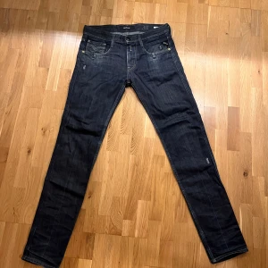Replay Anbass mörkblå jeans herr - Snygga mörkblå Replay Anbass jeans med klassisk femficksdesign och diskreta slitningar. Jeansen har raka ben, normal passform och är tillverkade i mjukt denimtyg med guldfärgade nitar och detaljer. Perfekta för en avslappnad och stilren look. Lätt använda, nyskick.