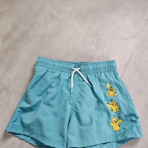 Turkosa Pikachu-shorts från H&M - Säljer ett par turkosa shorts från H&M med tre Pikachu-figurer tryckta på ena benet. Shortsen har vit snörning i midjan och är gjorda i lätt polyester, perfekta för chill dagar eller bad. Elastisk midja för skön passform. Är storlek 134/140 men upplever dem är små ,  tycker dem är som 122/128. Är i nyskick alldrig använda, då min son inte kunde ha dem.