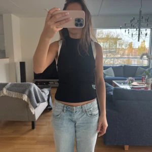 Svart ribbad polotopp från Maje - Säljer en svart ribbad polotopp från Maje med ärmlös design och hög hals. Toppen har en slim passform och är gjord i ett stretchigt material som sitter snyggt mot kroppen. Perfekt att styla med jeans eller kjol för en cool look.