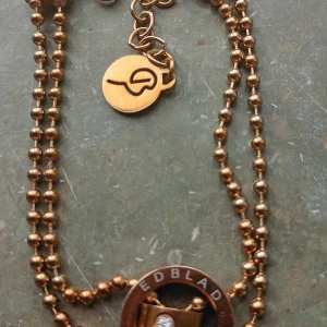 Guldfärgat halsband från Edblad - Snyggt guldfärgat halsband från Edblad med dubbla kulkedjor och en rund berlock med logotyp. Halsbandet har en cirkel med Edblad-text och en liten glittrande sten i mitten. Perfekt för dig som gillar stilrena detaljer.