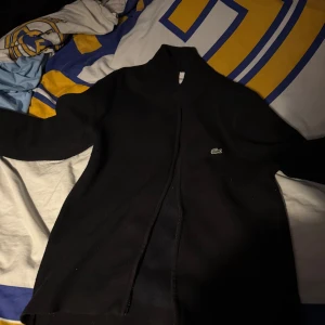 Svart cardigan jacka från Lacoste - Svart cardigan jacka från Lacoste med klassisk krokodillogga på bröstet. Jackan har dragkedja framtill och är långärmad med en stilren, enkel design. Perfekt för dig som gillar cleana och sportiga plagg med premiumkänsla.
