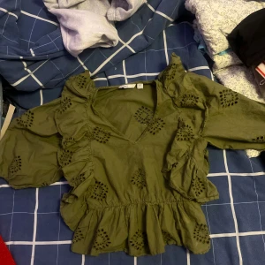 Grön blus med volanger från ONLY - Säljer en olivgrön blus från ONLY i storlek XS. Blusen har v-ringning, volangdetaljer längs axlar och framstycke samt broderade hålmönster över hela plagget. 