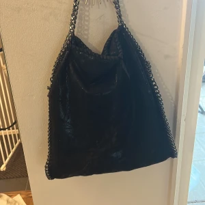 Svart axelväska med kedjedetaljer - Snygg svart axelväska med grova silverfärgade kedjor längs kanterna och som axelrem. Väskan har en mjuk, lite glansig yta och rymlig form, perfekt för att få plats med allt du behöver. Kedjorna ger en cool och edgy vibe.