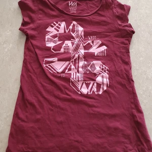 T-shirt - Säljer en vinröd t-shirt från WA Action Sports med coolt grafiskt tryck i ljusrosa på framsidan. T-shirten har rund hals och är kortärmad, perfekt för dig som gillar sportig och avslappnad stil. Materialet är mjuk bomull som känns skönt mot huden. Har ett litet hål.