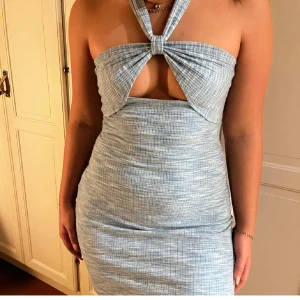 Blå kort ribbad klänning med knyt - Supersnygg blå kort klänning med ribbat tyg och diskret rutigt mönster. Halterneck med knyt i nacken och en cool cut-out framtill. Tight passform som framhäver figuren och ger en trendig look. Perfekt för dig som vill sticka ut med en unik detalj vid halsen.