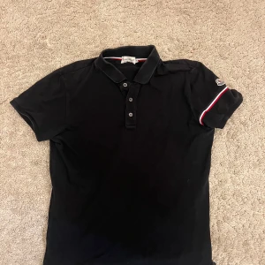 Svart Moncler pikétröja med röd/vit detalj - Svart pikétröja från Moncler i storlek S. Klassisk krage och tre knappar framtill. Ärmarna har snygga rödvita ränder och Moncler-logga på vänster arm. Jag vet inte om tröjan är äkta pga av den är köpt secondhand med lappen av klippt men om någon kunnig kan se om den är äkta via loggan där av de låga priset. Hör av er vid frågor 
